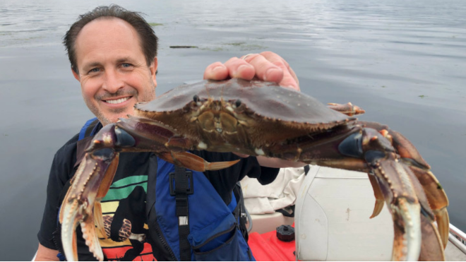A man on a boat shows off a big crab he just caught. He's smiling and wearing a life vest.