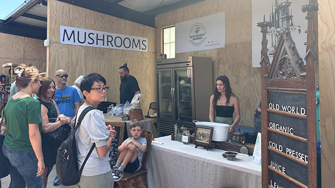 Several people stand by a table at a local farmers market. To the left-hand side is a sign that says "Mushrooms." There is a vendor behind the table and to the right is a sign that says, "Old world organic fresh pressed juice."