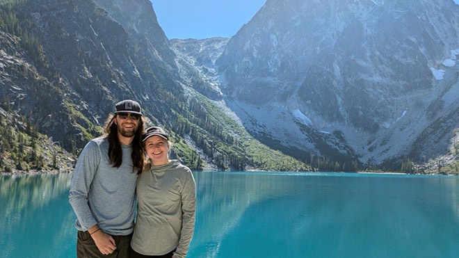 A couple poses in front of a lake, with mountains reflecting in the bright blue water behind them.