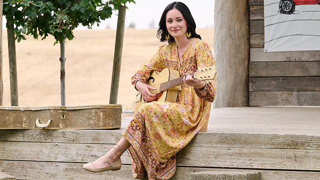 BECU member Annie M. sitting on a wooden porch, wearing a yellow dress, playing guitar with legs crossed.