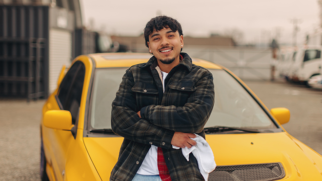 BECU member Santos G. stands in front of a yellow car, smiling with arms crossed in an outdoor auto shop setting