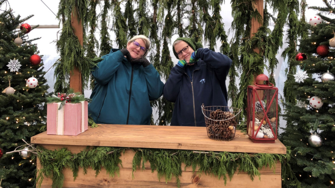 Two women pose for a festive photo at a booth at Christmas Tree farm. They are both wearing gloves and a hat.