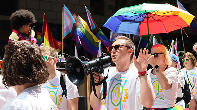 A BECU employee speaks into a megaphone while participating in the Seattle Pride Parade. A woman carries a rainbow umbrella in the background. A DJ and Pride flags are also in the background.