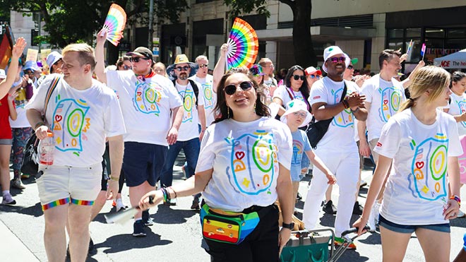 BECU employees wearing matching tee shirts march in the Seattle Pride Parade. A smiling woman is at the center of the photo. People smile and wave rainbow fans in the background.