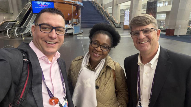 Three professionals wearing conference badges pose together for a selfie inside a large convention center, with escalators and a wide staircase visible in the background.