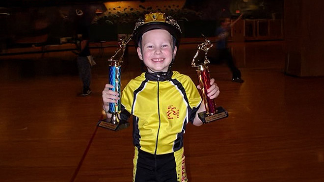 Young Cooper McLeod holding two skating trophies while wearing a yellow racing outfit and a helmet. 