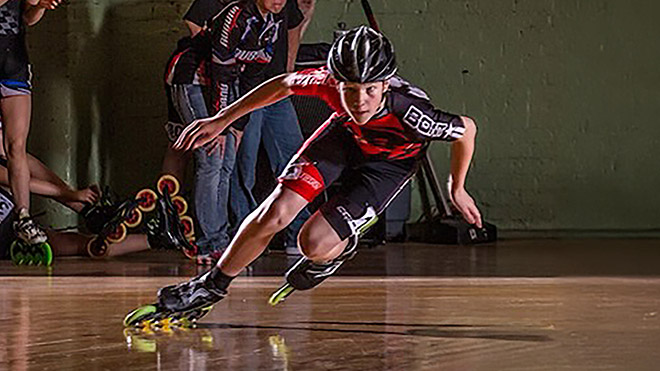 Teenage Cooper McLeod in a red and black racing suit taking a sharp turn on an indoor roller rink as others watch.