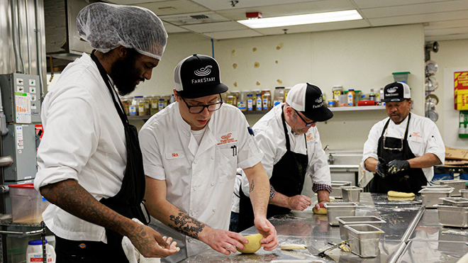 Four cooks prepare food at a stainless-steel counter in a commercial kitchen.