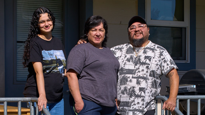 Three people stand together outside a building, smiling at the camera.