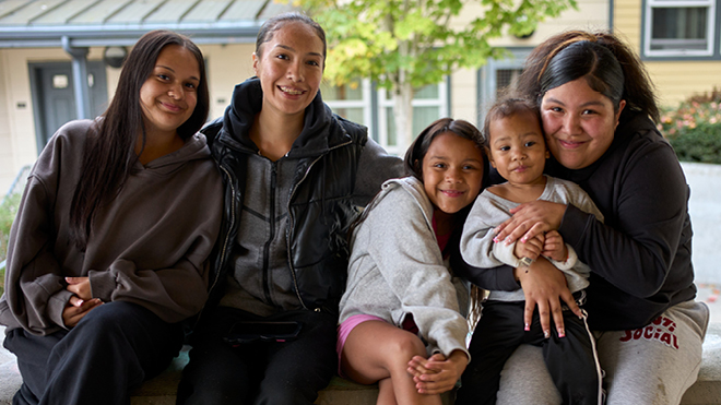 Four smiling adults and children sit closely together outdoors, with one person holding a toddler.