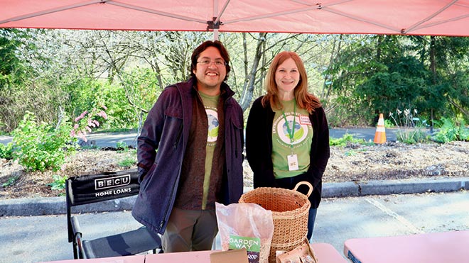 Two BECU employees wearing spring green T-shirts with the Environmental Sustainability Employee Resource Group logo on the front smile while standing under a red tent.