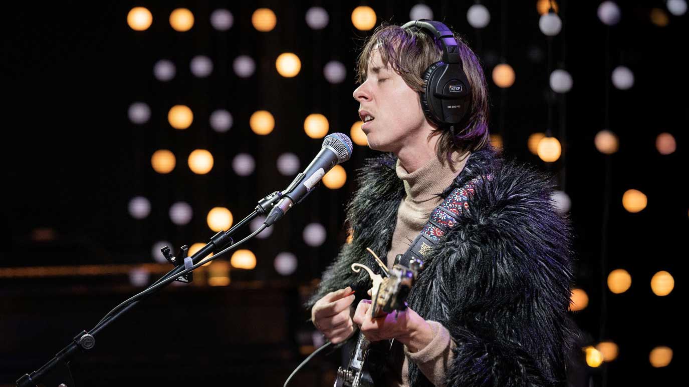 A musician wearing headphones performs with an electric guitar at a microphone, with stage lights twinkling in the background.