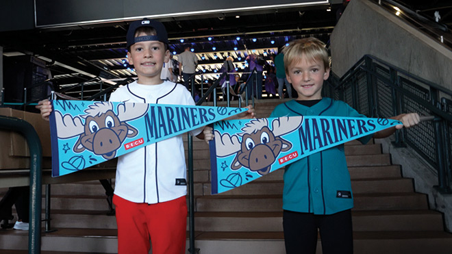 Two boys standing inside T-Mobile stadium wear Seattle Mariners jerseys and hold Mariners pennants with the Mariner Moose and BECU logo.