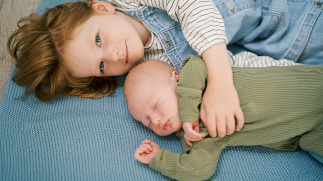 An older child lies on a blue mat with an arm gently around a newborn dressed in green, resting side by side.
