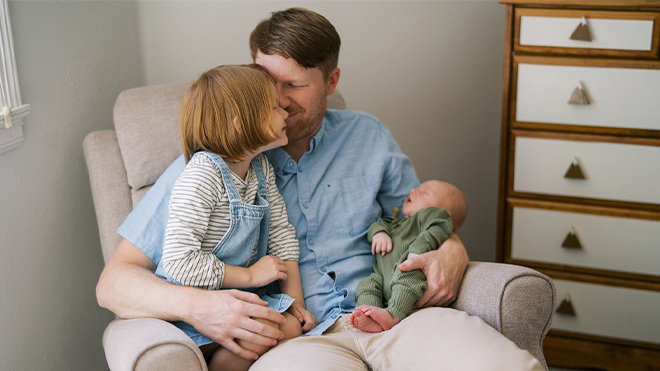 An adult sits in an armchair holding a newborn while a young child sits close and looks toward the baby.