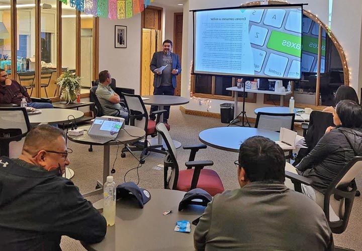 A group of about six individuals are seated at round tables in a classroom setting, while one person is standing and presenting in front of the group. The person presenting is holding a piece of paper and talking. The presenter is standing in front of a projection screen. Additionally, there are multi-colored flags hanging from the ceiling. 