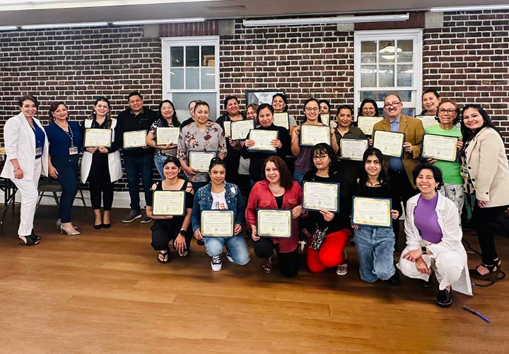 A large group of more than 20 people are standing and kneeling inside in front of a brick wall, while many of them hold up financial literacy certificates.
