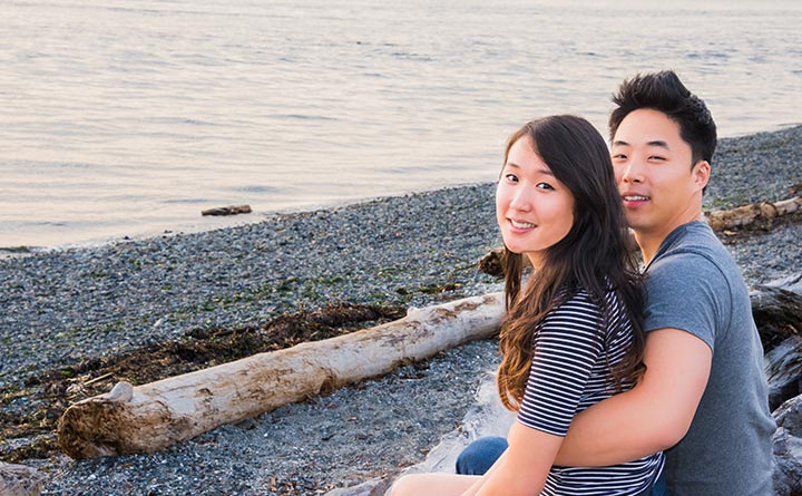 A man and woman sit together on a log near the water. The beach is a combination of sand and rocks. The water is calm. The man has his arm around the woman. They are looking over their shoulders to smile at the camera.
