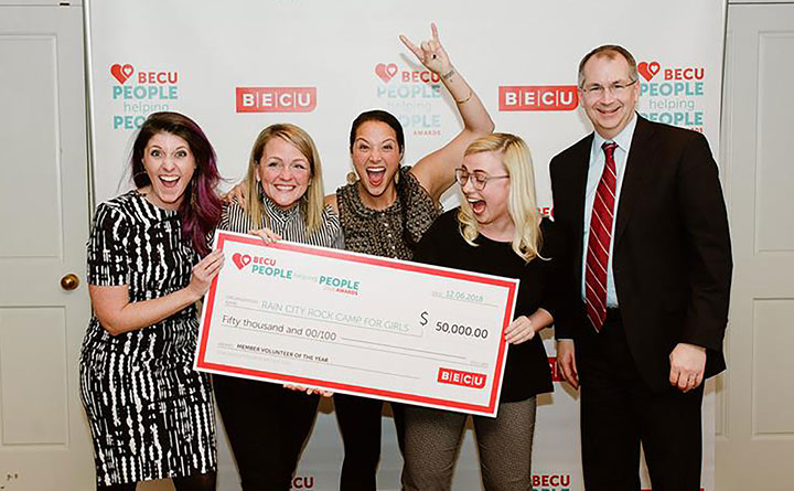 Benson stands with four women holding a giant check for $50,000 in front of a backdrop that says BECU People Helping People Awards. The check is to Rain City Rock Camp for Girls. The women have big smiles, and one woman is doing the rock-and-roll salute, holding her hand up to show horns with her index and little finger.