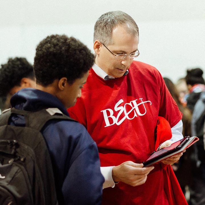 Benson Porter, wearing a red T-shirt with the BECU logo across the front, leans over to look at a tablet screen with a student. 