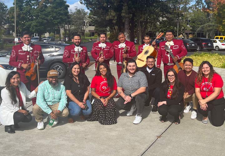 Nine BECU employees kneel in front of a six-member mariachi band outside of BECU's headquarters in Tukwila. Several employees are wearing red Latinx Employee Resource Group (ERG) t-shirts. The mariachi band members are holding instruments like trumpets, guitars and violins. In the background there are trees and cars.