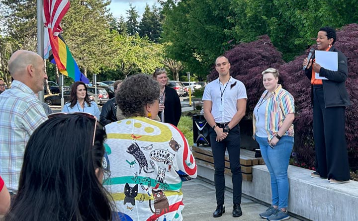 BECU CEO Beverly Anderson holds a microphone to address a gathering of BECU employees standing outside of Tukwila Financial Center prior to a Pride flag raising. A flag pole with a lowered U.S. flag and Pride flag is in the background.