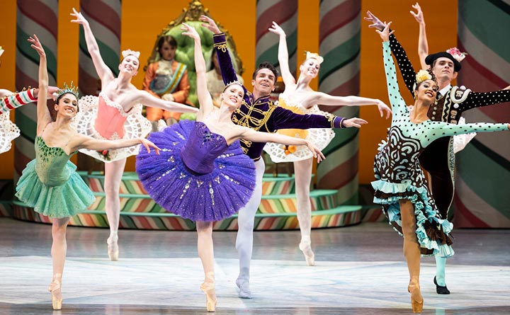 In this photo ballet dancers are performing on a stage. The photo depicts a scene from the Nutcracker ballet. There are seven people in the image and each dancer is posing on one leg while raising their hands in the air.