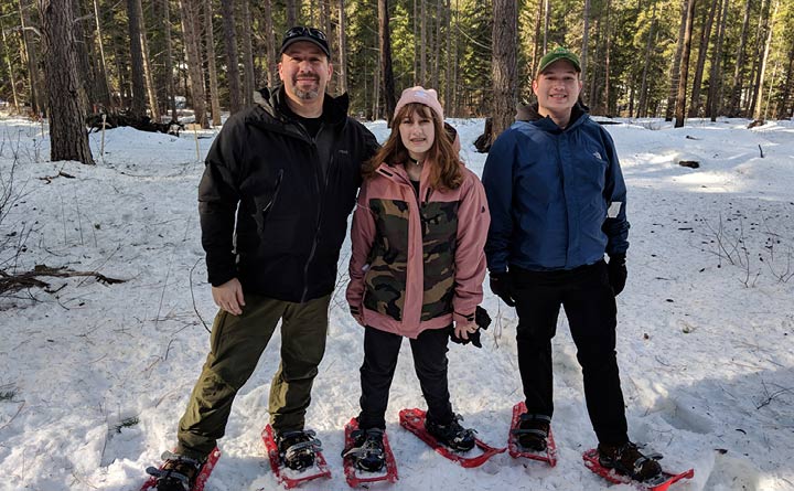 A family of three poses for an outdoor photo while snowshoeing. There is snow on the ground. All three of the people in the photo have snowshoes on their feet. Each person is bundled in winter clothes. Behind them are trees and snow.
