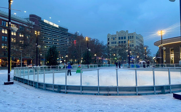 This is an image of an outdoor ice skating rink. In the photo two children are ice skating. There are city buildings in the background and it's a dusk evening. 