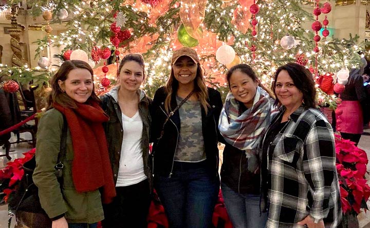 Five people pose indoors in front of a large holiday tree. The tree has many lights and decorations on it. The tree is very colorful and had red, white and green ornaments. Two of the people who are posed in front of the photo are wearing large scarves. All five of the people in the photo are standing and smiling. 