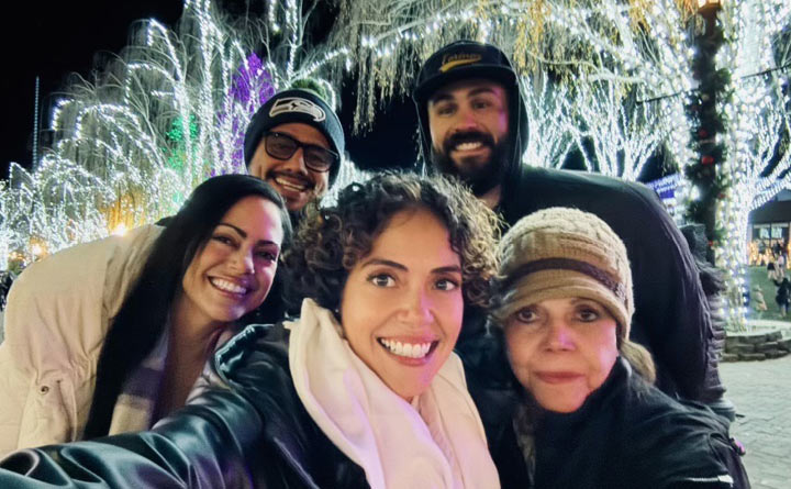 Five people pose for a selfie in front of a lighted holiday trees in Leavenworth, Washington. The five people are all smiling and dressed in winter clothing such as beanies, hats and scarves. There is snow on the ground and it's night time outside. 