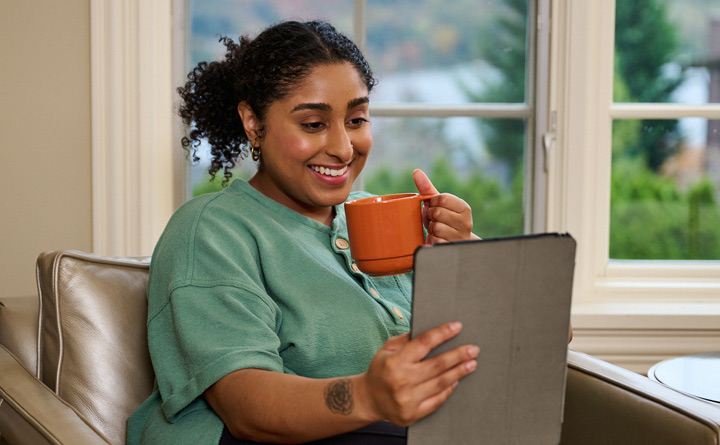 Young person smiling and holding a coffee cup while looking into a mobile tablet 