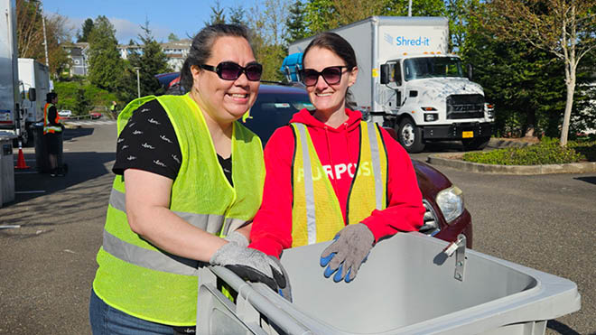 Two smiling workers wearing sunglasses and safety vests with a shred bin.