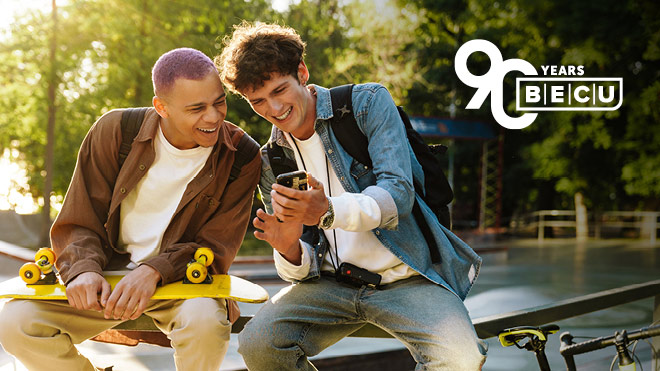 Two smiling men outdoors, one is holding a skateboard, and both men are looking at a mobile phone
